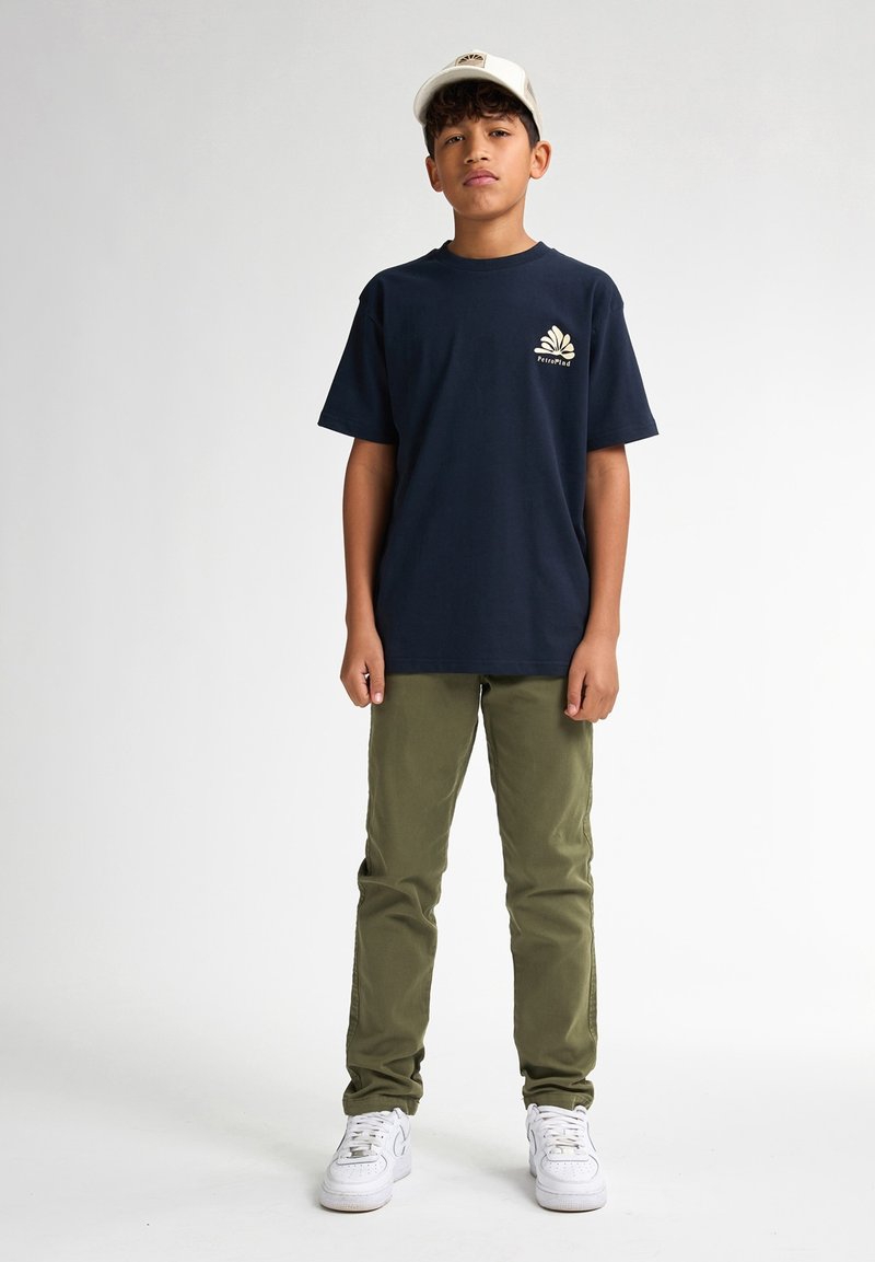 Marineblauwe katoenen t-shirt met een wit logo op de borst, gecombineerd met groene katoenen broek en witte sneakers. Een eenvoudige pet maakt de outfit compleet.