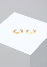Gold-tone adjustable cuff rings, featuring a smooth surface on one and a textured design on the other, displayed on a white pedestal.