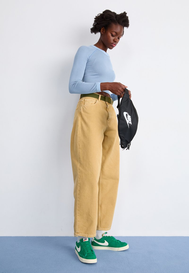 Camiseta de manga larga de rayas azul claro, pantalones de denim beige holgados, cinturón verde y zapatillas Nike verdes. Sosteniendo una bolsa negra de Nike con el logo.