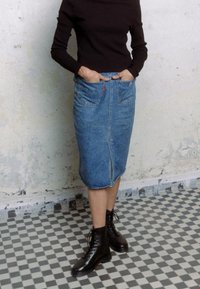 Falda lápiz de mezclilla con abertura delantera, lavado azul medio, dos bolsillos frontales, combinada con botines negros de cordones y una blusa negra ajustada.