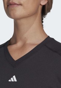 Camicia nera con scollo a V realizzata in tessuto liscio, con un logo bianco. Primo piano del collo e del busto superiore, che mostra i dettagli delle cuciture.