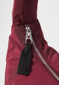Burgundská nylonová taška se stříbrným zipem a černou textilní stahovací šňůrkou, která má integrovanou logo nálepku v designu zipu.