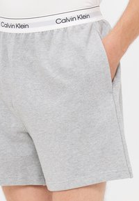 Graue Baumwollshorts mit lockerer Passform, ausgestattet mit seitlichen Taschen und einem elastischen Bund, auf dem "Calvin Klein" in schwarzem Text zu sehen ist.