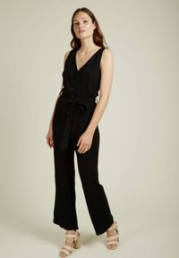 Combinaison sans manches noire avec un col en V, fermeture à boutons, taille ceinturée et jambes larges. Complétée par des sandales à talons beiges.