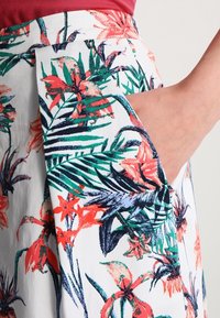 Jupe à poches en tissu blanc ornée de motifs tropicaux vibrants en vert, rouge et bleu. Texture douce avec une coupe décontractée.