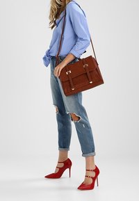 Ruskea mokkahan handbag kultaisilla yksityiskohdilla, suorakaiteen muotoinen ja kaksi kahvaa, yhdistettyinä revittyihin farkkuihin ja punaisiin korkokenkiin.