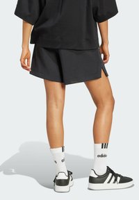 Čierne športové šortky so bočnými rozparkami, skombinované s čierno-bielymi teniskami a pruhovanými bielymi ponožkami. Materiál vyzerá hladko, s neformálnym dizajnom.