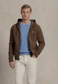 Hoodie castanho com fecho éclair e cordões, sobre um suéter de malha azul claro, combinado com calças de cor creme.