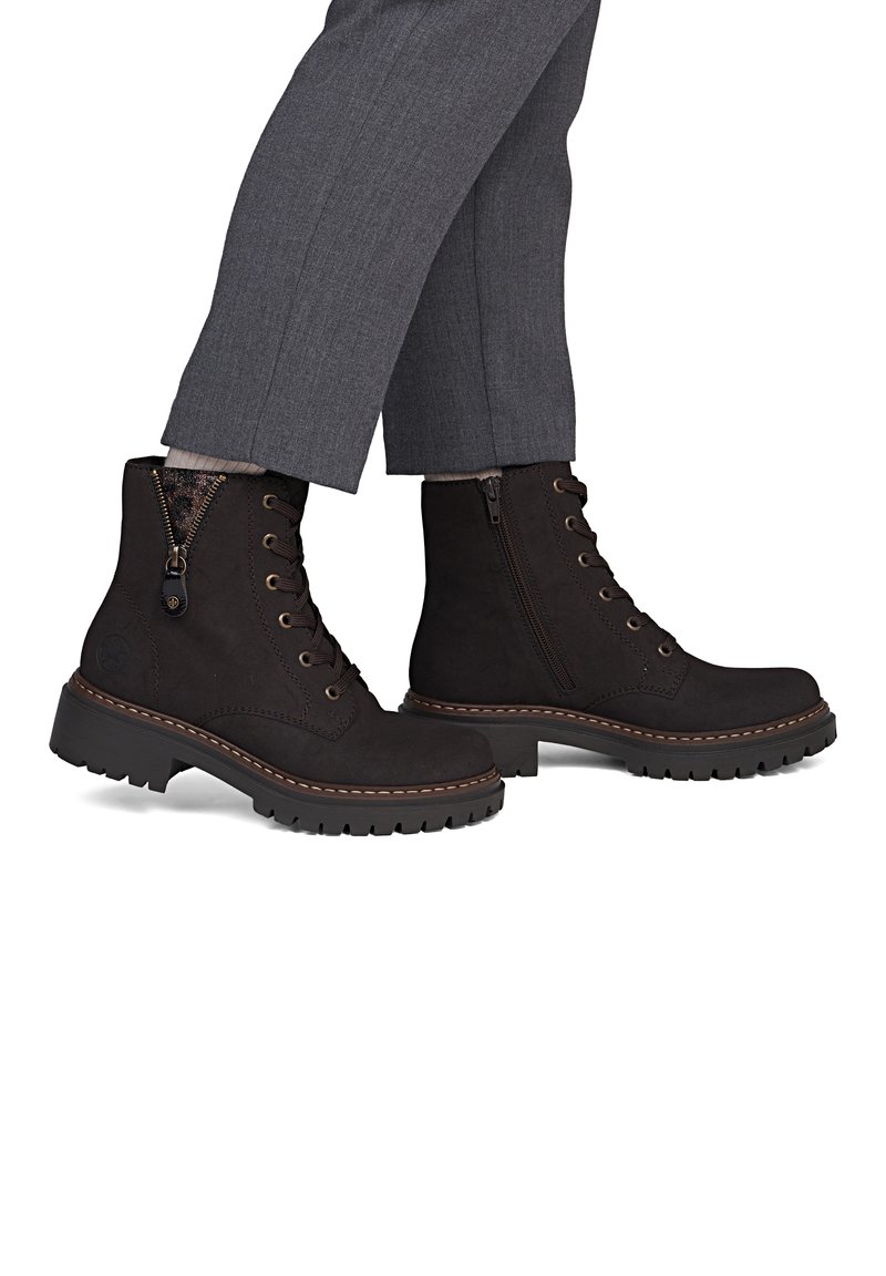 Bottines marron foncé à lacets avec fermetures éclair latérales portées avec un pantalon gris, montrant des semelles robustes et des coutures contrastées sur fond blanc.