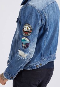 Džínová bunda s roztrženým detailem na rukávu a dvěma nášivkami s motivy z přírody, nošená přes černé kalhoty na osobě s tetovanou rukou.