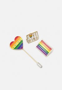 Três pins de esmalte: um coração com "PRIDE" em riscas arco-íris, "LOVE WINS" com um coração, e uma bandeira arco-íris, todos com detalhes em dourado.