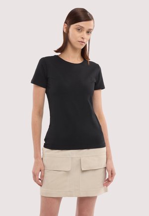 Młoda kobieta o prostych brązowych włosach ubrana w dopasowaną czarną koszulkę z krótkim rękawem oraz beżową mini spódnicę z dużymi przednimi kieszeniami.