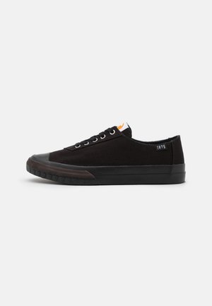 Zapatillas de lona negras con suela de goma, cuentan con ojales blancos, una puntera negra y un pequeño acento naranja en la lengüeta. Diseño minimalista.