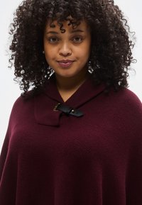 Bordový pletený pončo so zloženým golierom, ozdobený čiernou koženou prackou a zlatými kovovými doplnkami. Mäkká textúra a pohodlný strih.