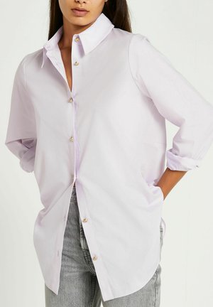 Vrouw draagt een losse lichtpaarse blouse met knopen en opgerolde mouwen en grijze spijkerbroek, handen in de zakken, tegen een effen achtergrond.