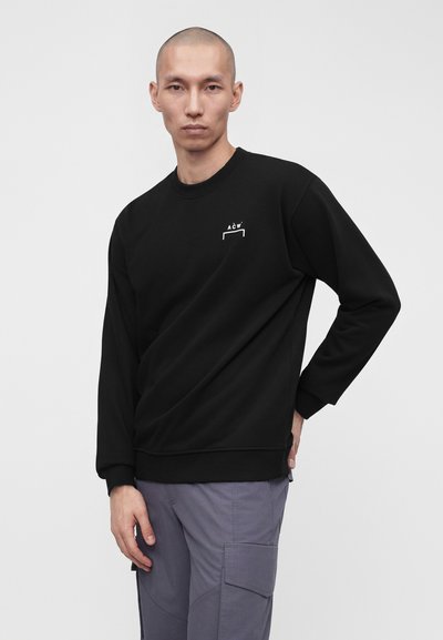 Άνδρας με μαύρο φούτερ μακρυμάνικο με λευκό λογότυπο ACW και γκρι παντελόνι cargo, στέκεται μπροστά σε απλό φόντο.