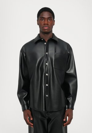 Man wearing a black faux leather button-up shirt paired with matching black faux leather pants, standing against a plain white background.