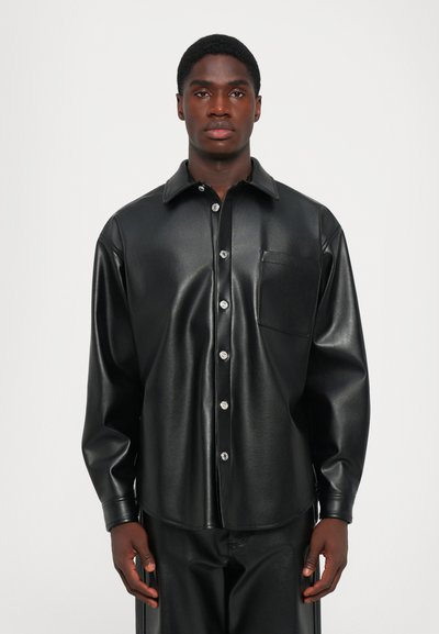 Uomo che indossa una camicia nera in finta pelle con bottoni abbinata a pantaloni neri in finta pelle, in piedi di fronte a uno sfondo bianco uniforme.