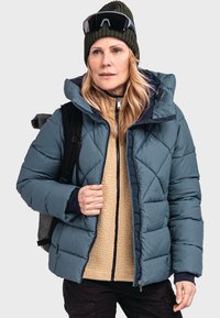 Türkisfarbene Puffjacke mit diamantartigem Steppmuster, ausgestattet mit einer abnehmbaren Kapuze, über einem beige-farbenen Fleece-Oberteil, mit schwarzen Akzenten und Reißverschlüssen.