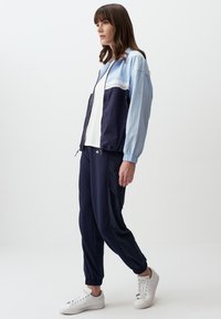 Giacca zip blu chiaro e navy con polsini elastici, abbinata a pantaloni jogger navy scuro e sneakers bianche. Tessuto liscio, design casual.