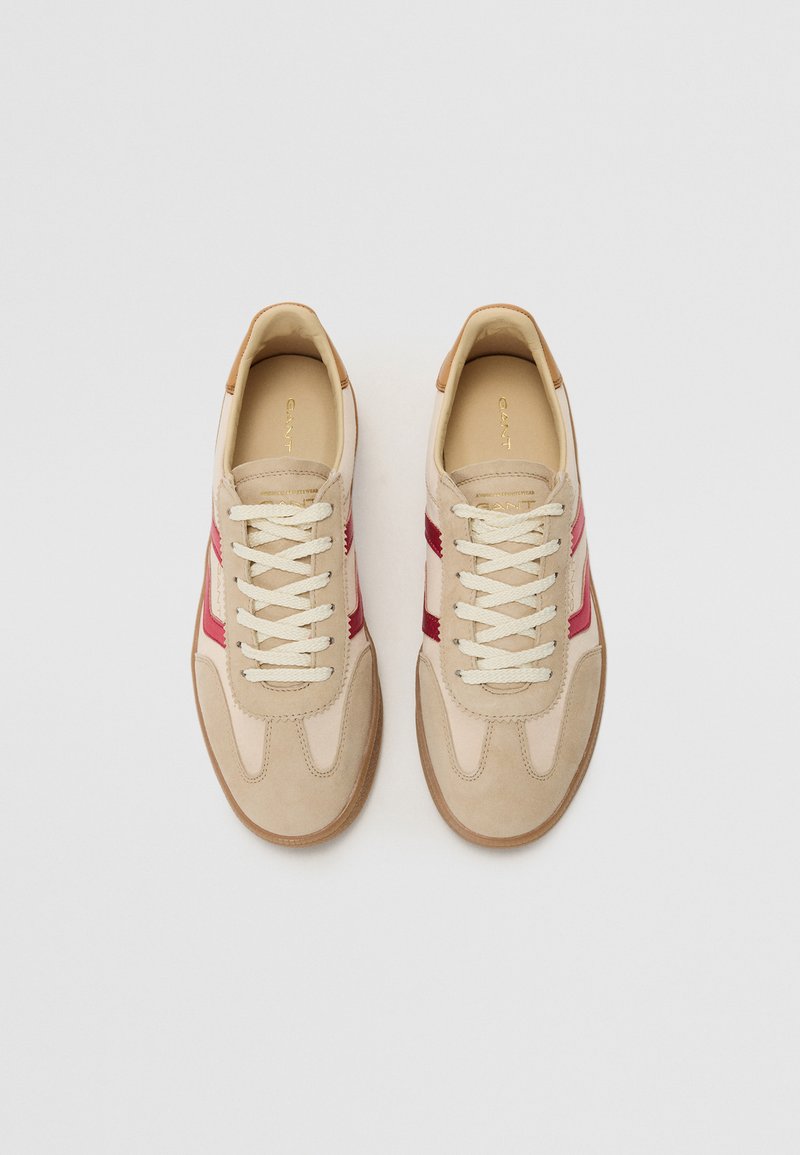 Beżowe sneakersy z czerwonymi akcentami, wykonane z zamszu i skóry, z okrągłym noskiem, płaskimi sznurowadłami i gumową podeszwą. Nazwa marki widoczna w środku.