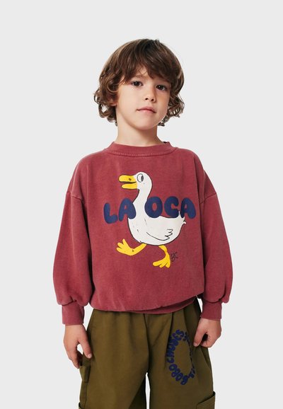 Felpa bordeaux con una grafica di anatra bianca con becco e zampe gialle, testo "LA OCA" in blu. Vestibilità ampia con polsini a costine.