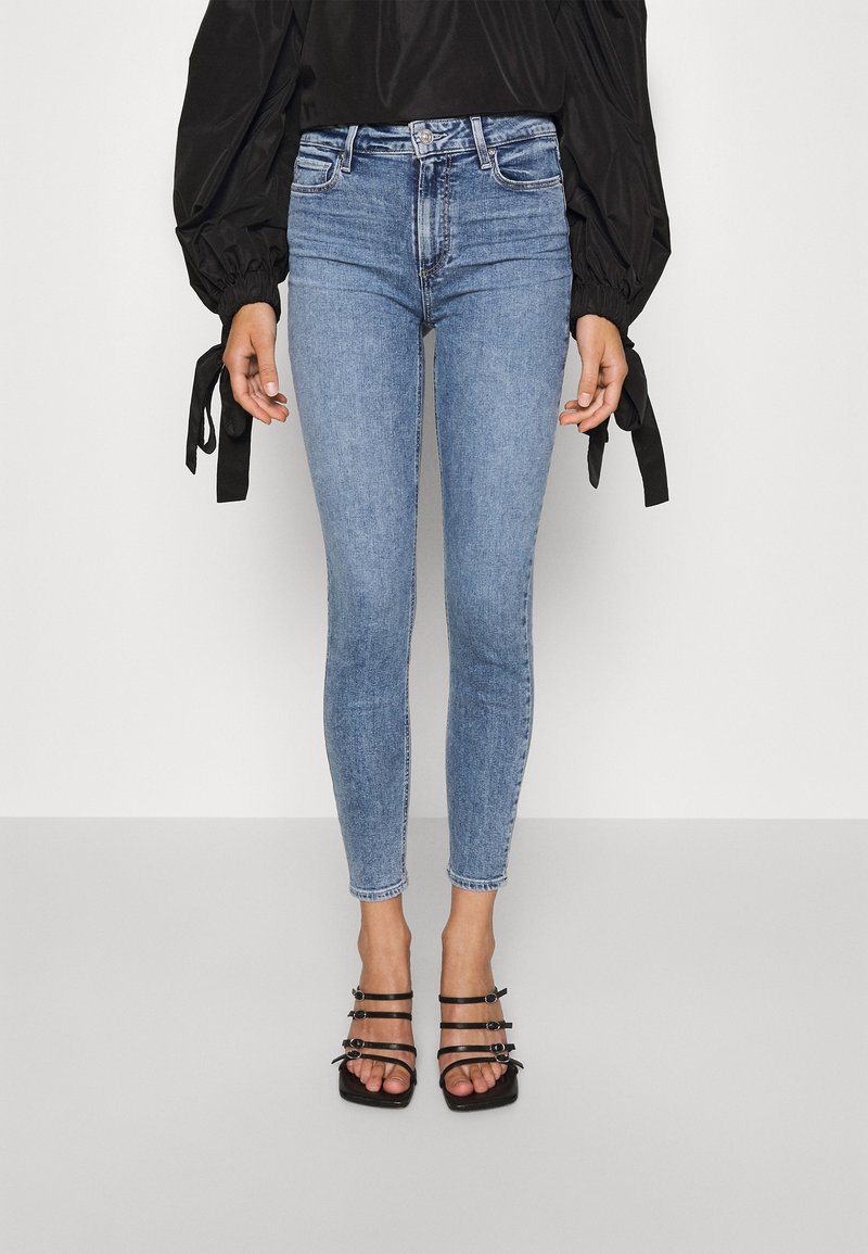 Jeans de mezclilla azul ajustados con una cintura alta, que muestran una silueta ceñida. Combinados con sandalias de tacón negro con tiras.