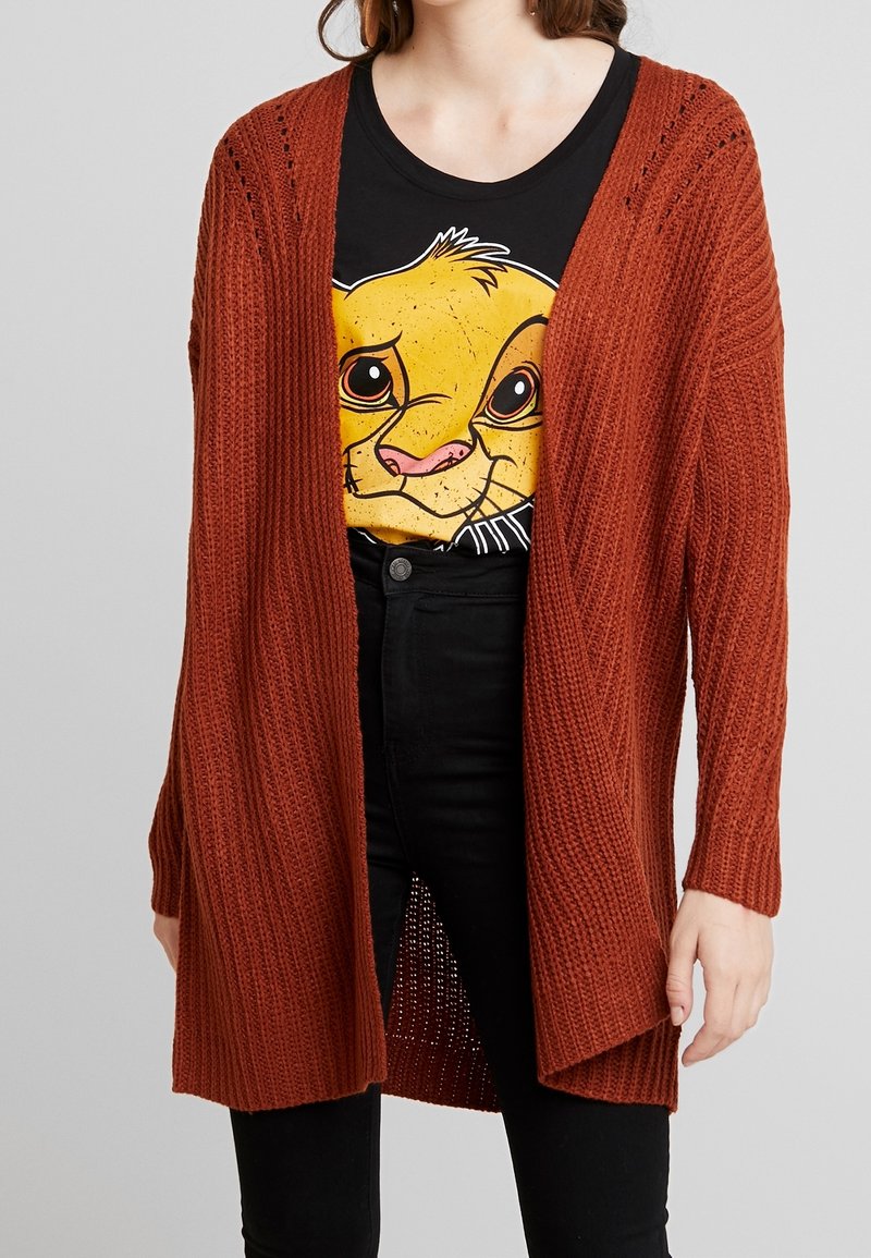 Cardigan couleur rouille avec texture côtelée, devant ouvert et manches longues. Porté sur un t-shirt noir avec un dessin d'animal de dessin animé.
