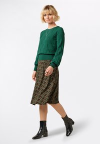 Grüner Pullover mit Blättermuster, knielanger Blumensatinrock in dunklen Farbtönen und schwarze lackierte Lederstiefeletten. Schlichte Halskette als Akzent.