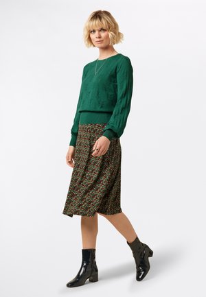 Suéter verde con patrón de hojas, falda floral de longitud media en tonos oscuros y botines de charol negros. Acento con un collar simple.