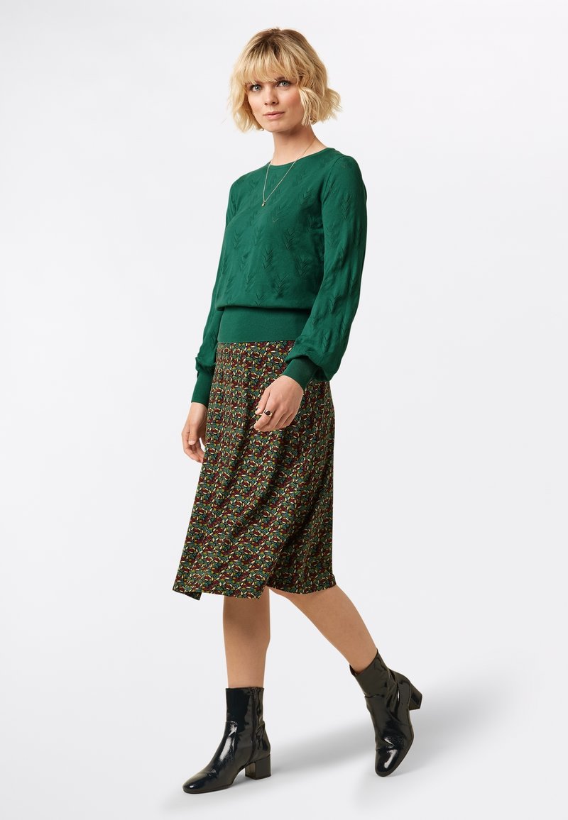 Grüner Pullover mit Blättermuster, knielanger Blumensatinrock in dunklen Farbtönen und schwarze lackierte Lederstiefeletten. Schlichte Halskette als Akzent.