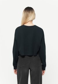 Haut noir cropped à manches longues avec un col rond et une texture lisse, associé à un pantalon gris foncé avec des poches arrière.