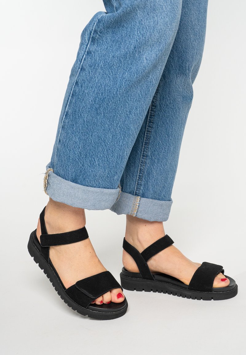 Fødder iført sorte sandaler med åbne tæer og justerbare remme, båret med opsmøgede blå jeans på en ens hvid baggrund.