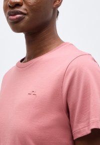 Κοντινό πλάνο ενός ατόμου που φοράει μια απλή ροζ κοντομάνικη μπλούζα με ένα μικρό ανάγλυφο λογότυπο στο στήθος, πάνω σε λευκό φόντο.