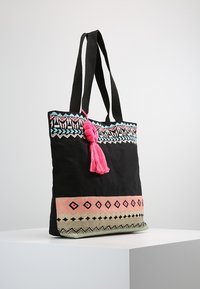 Bolsa de tela negra con ribete de patrón colorido. Presenta un borlas rosa, diseños geométricos en turquesa, rosa y verde. Correas duraderas.