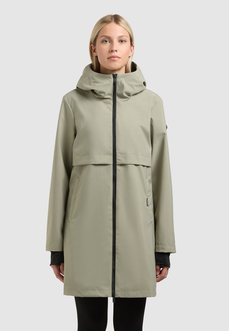 Jeune femme portant un imperméable à capuche gris clair avec une fermeture éclair frontale, debout devant un fond clair uni.