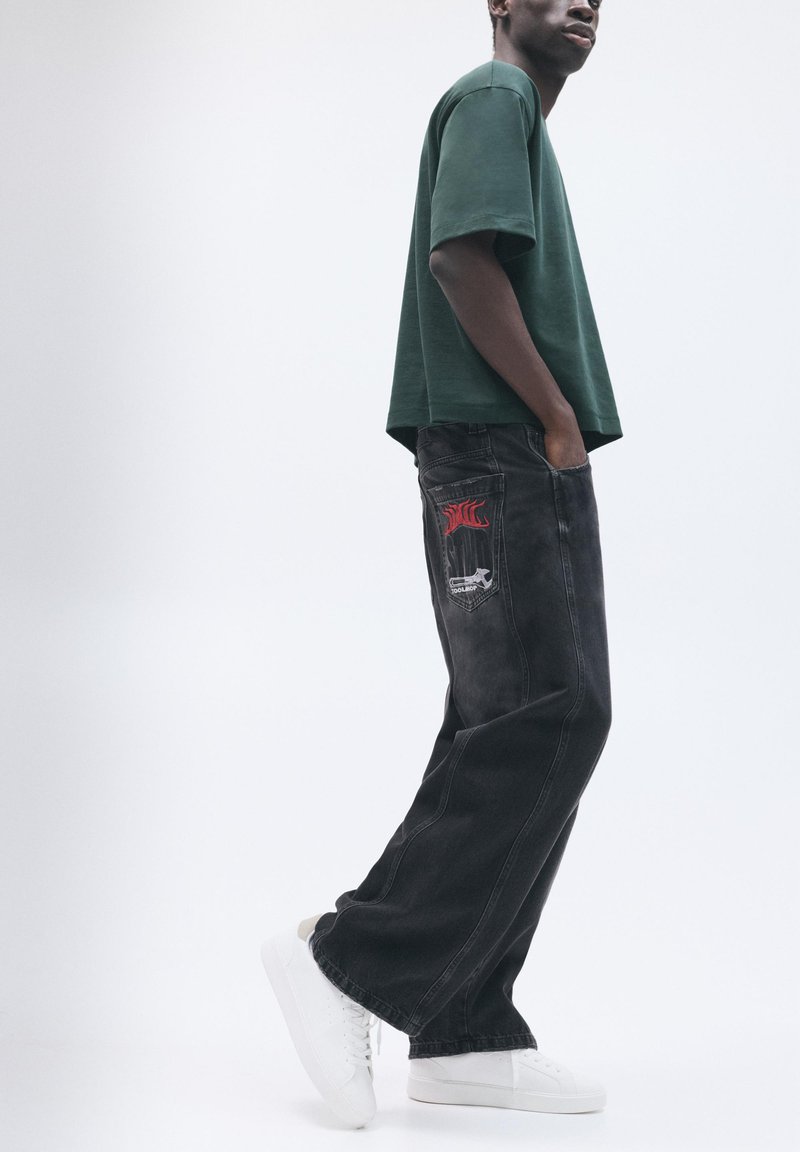 Mann steht mit Händen in den Taschen, trägt ein dunkelgrünes Hemd, schwarze Jeans mit grafischem Aufnäher und weiße Sneakers auf weißem Hintergrund.