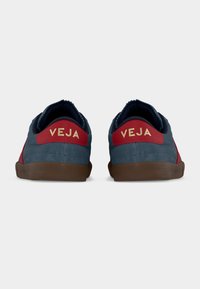 Ζευγάρι από σκούρα μπλε και κόκκινα αθλητικά παπούτσια με καφέ σόλες, εμφανιζόμενα από πίσω, με την επωνυμία «VEJA» σε μπεζ γράμματα.