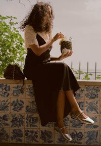 Vrouw met krullend haar in een zwarte jurk en kanten top zit op een betegelde rand en houdt een ananasdrank met rietjes en een schijfje citroen vast.