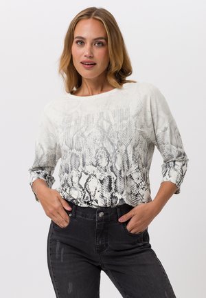 Frau trägt ein weiß-grau schlangenmusterlangärmeliges Oberteil und schwarze Jeans, steht mit den Händen in den Taschen vor einfachem Hintergrund.