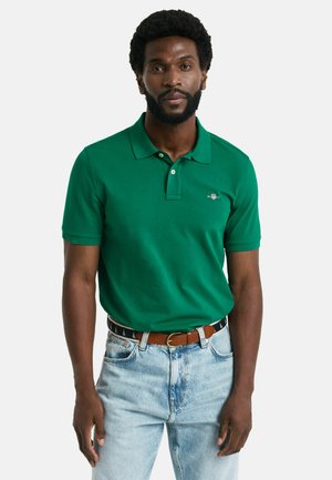 Green polo shirt made of cotton, featuring a two-button placket and logo on the chest, paired with light denim jeans and a brown belt.