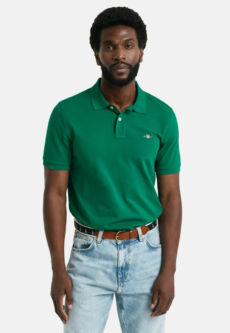 Grünes Poloshirt aus Baumwolle mit einer Knopfleiste mit zwei Knöpfen und einem Logo auf der Brust, kombiniert mit hellblauen Jeans und einem braunen Gürtel.