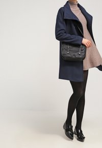 Cappotto di lana navy, vestito aderente grigio a collo alto, borsa a tracolla nera, collant neri e scarpe basse. Design semplice, vestibilità sartoriale, hardware minimale.