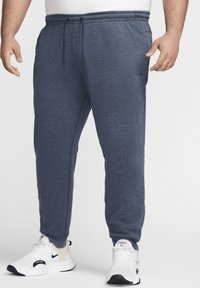 Pantalones deportivos azul marino con cintura elástica y cordón, bolsillos laterales, piernas ajustadas y logo de Nike en la cadera izquierda. Llevados con zapatillas blancas.