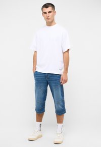 Weißes T-Shirt mit kurzen Ärmeln, blaue Denim-Cropped-Hosen mit Stickdetails, cremefarbene Sneaker, gemusterte weiße Socken. Einfaches, lässiges Outfit.