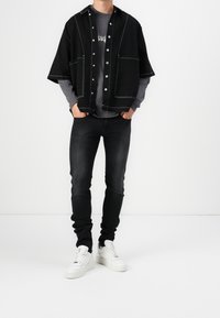 Camiseta negra corta con costuras blancas sobre una camiseta de manga larga gris, combinada con jeans ajustados negros y zapatillas blancas.