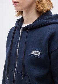 Νεαρό άτομο με κοντά ξανθά μαλλιά, που φοράει μια μπλε σκούρα ζακέτα με φερμουάρ και μαύρο κορδόνι, με ένα λογότυπο Nørgaard.