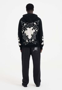 Sweat à capuche noir avec un design graphique blanc, présentant un cœur et des os. Associé à un pantalon gris foncé ample.