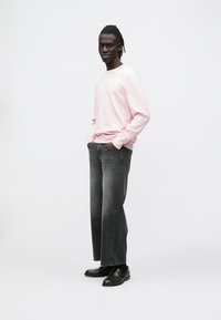 Šviesiai rožinis džemperis laisvo kirpimo, derinamas su tamsiai pilkais siaurėjančiais džinsais ir juodais loaferiais. Paprastas dizainas, be matomų raštų ar logotipų.