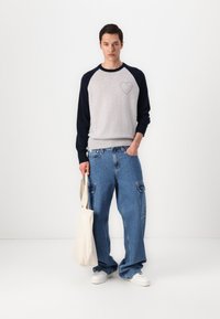 Gray sweatshirt with navy raglan sleeves, featuring a small heart detail. Loose-fitting blue cargo jeans and a cream tote bag. White sneakers.